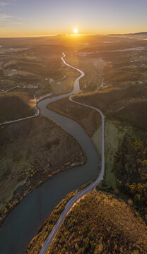 Aerial view of the winding Seixe river flowing through the lush landscape at sunset, with golden light reflecting on the water and surrounding hills in Odeceixe, Portugal.