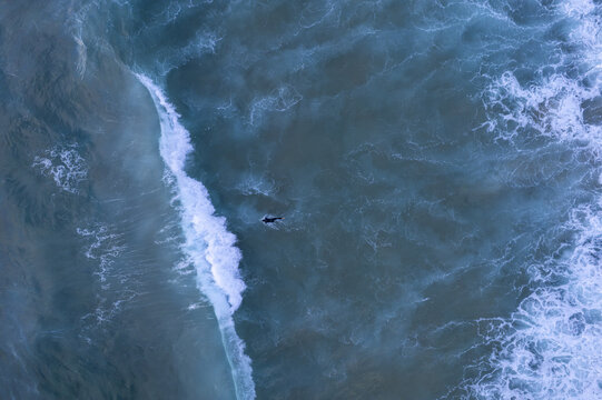 Aerial view of a lone surfer paddling in the deep blue ocean with white breaking waves near the coast of Sagres, Faro, Portugal.