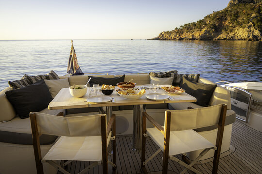 Mallorca, Spain - 20 April 2024: View of a dining table set on the deck of a yacht with appetizers and drinks, overlooking the Mediterranean Sea and rocky cliffs during golden hour.