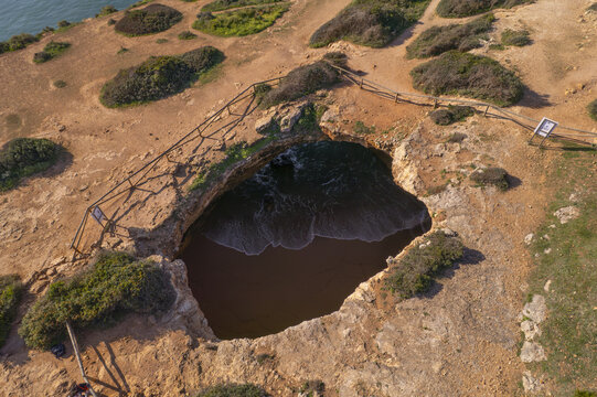 Aerial view of Benagil Cave, featuring a natural sea cave opening with a sandy beach inside, surrounded by rugged coastal cliffs and wooden safety railings in Lagoa, Faro District, Portugal.