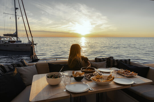 Balearic Islands, Spain - 20 April 2024: View of a woman sitting at a table with food on a sailboat during sunset in Mallorca.