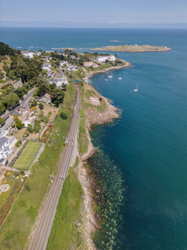 Aerial view of the coastal railway line passing through Dalkey with Dalkey Island in the background, featuring rocky cliffs, residential houses, and the Irish Sea in Dublin, County Dublin, Ireland.