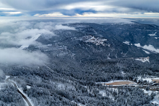 Aerial view of the snow-covered Mummelsee area in the Black Forest with dense pine forests, winding roads, and low-hanging clouds in Seebach, Baden-Wurttemberg, Germany.
