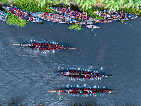 Gopalganj, Dhaka Division, Bangladesh - 30 October 2023: Aerial view of traditional long boats filled with people rowing along a river, surrounded by lush green vegetation.