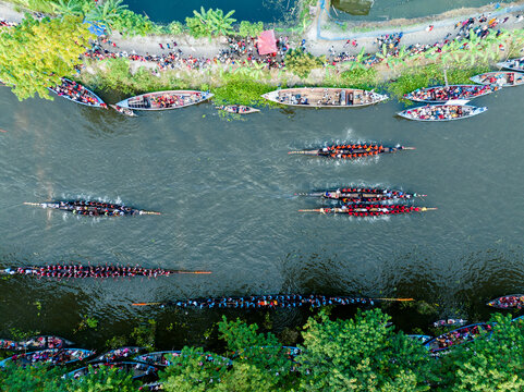 Gopalganj, Bangladesh - 30 October 2023: Aerial view of a traditional boat race on the river with many long wooden boats filled with rowers and spectators gathered along the lush green banks.