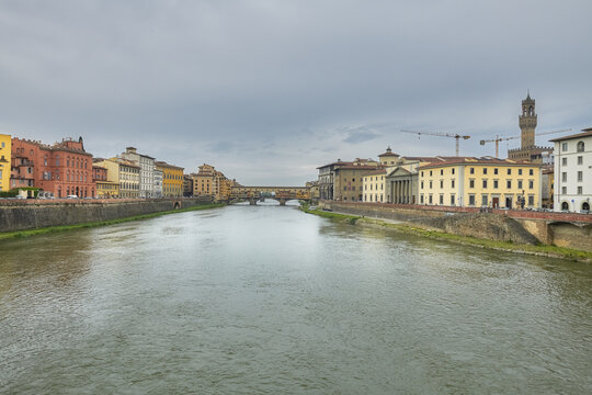 Florence, Italy - 05 May 2022: View of the Ponte Vecchio bridge spanning the Arno River with historic buildings and the Palazzo Vecchio tower in the background under a cloudy sky.