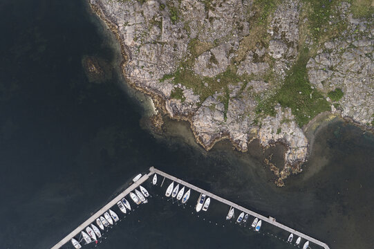 Aerial view of Sando marina with docked sailboats along a wooden pier, rocky coastline, and dark water in Vallda, Hallands lan, Sweden.