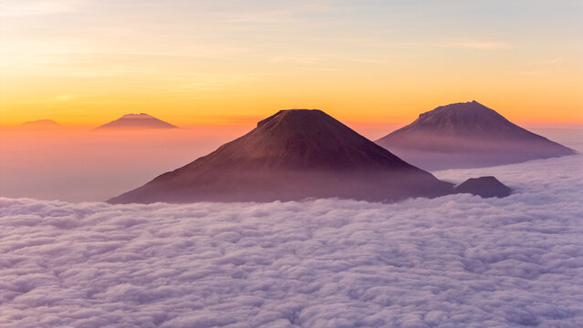 Aerial view of Mount Bismo and surrounding volcanic peaks rising above a thick sea of clouds during a vibrant golden hour sunrise in Kecamatan Kejajar, Jawa Tengah, Indonesia.