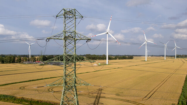 Aerial view of wind turbines in a golden wheat field with a large electricity pylon in the foreground under a blue sky in Gdansk, Pomeranian Voivodeship, Poland.
