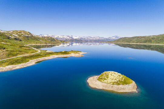 Aerial view of the calm blue waters of Lake Tyin with a small grassy island, surrounded by rugged mountains and a winding road in Tyinkrysset, Innlandet, Norway.
