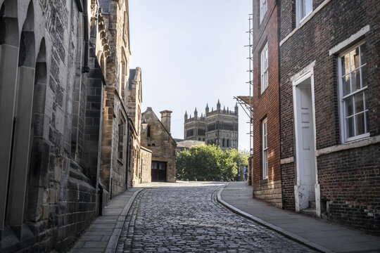 View of the historic Durham Cathedral towers rising above a narrow cobblestone street lined with traditional stone and brick architecture in Durham, England, United Kingdom.