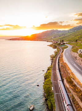 Aerial view of a passenger train traveling along the scenic shoreline of Lake Geneva, passing through the terraced vineyards of Lavaux at sunset in Bourg-en-Lavaux, Vaud, Switzerland.