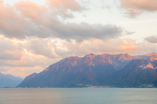 View of Lake Geneva with the Alps in the background during golden hour, featuring soft pink and orange clouds over the mountains in Bourg-en-Lavaux, Vaud, Switzerland.