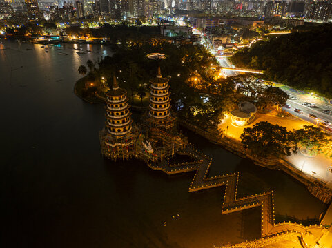 Aerial view of the illuminated Dragon and Tiger Pagodas at Lotus Pond with a zigzag bridge, surrounding parkland, and city lights at night in Kaohsiung, Taiwan.
