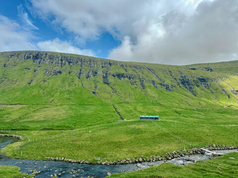 View of a small green building nestled in the lush grassy hills of the Faroe Islands with a stream flowing through the foreground under a cloudy sky, Faroe Islands.