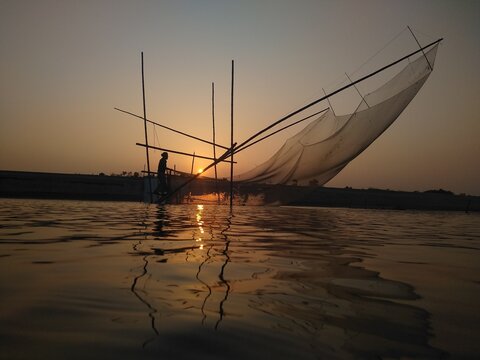 View of a fisherman using a traditional lift net on the river at sunset in Kushtia, Khulna Division, Bangladesh.