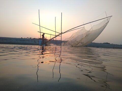View of a fisherman operating a traditional lift net in the river at sunset with calm water reflections in Kushtia, Khulna Division, Bangladesh.