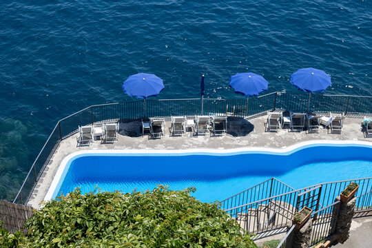 Aerial view of a swimming pool with blue umbrellas and lounge chairs overlooking the sea in Amalfi, Campania, Italy.