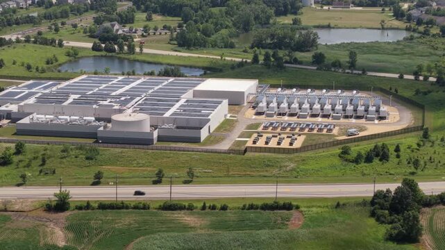 Aerial view of a modern data center facility featuring solar panels and cooling units, surrounded by green fields and a pond in Volo, Illinois, United States.