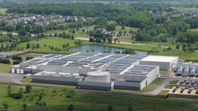 Aerial view of a large data center facility with solar panels on the roof, surrounded by green fields and a small pond in Volo, Illinois, United States.