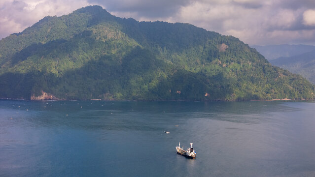 Aerial view of Pantai Karanggongso featuring a cargo ship sailing in the calm blue bay near lush green mountains under a cloudy sky Watulimo, East Java, Indonesia.