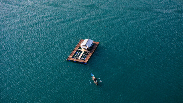 Aerial view of Pantai Karanggongso featuring a floating fish farm structure and a small wooden boat on the calm blue sea in Watulimo, East Java, Indonesia.