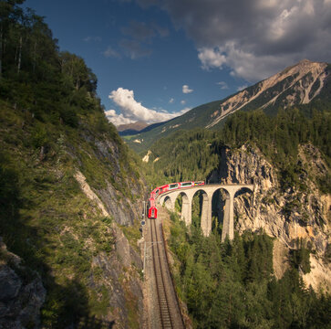 Aerial view of the Landwasser Viaduct with a red train crossing the stone arches in a steep mountain valley surrounded by pine trees in Bergun Filisur, Graubunden, Switzerland.