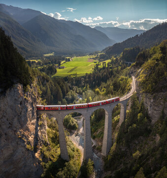 Aerial view of the Landwasser Viaduct with a red train crossing the stone arches, set against alpine forests, mountains, and a river valley in Bergun Filisur, Graubunden, Switzerland.