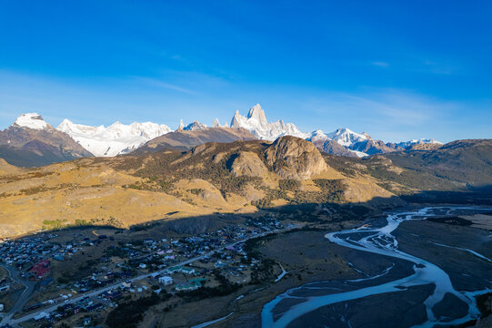 Aerial view of El Chalten nestled in the Patagonian landscape with snow-capped peaks of Mount Fitz Roy in the background under a clear blue sky, El Chalten, Santa Cruz, Argentina.