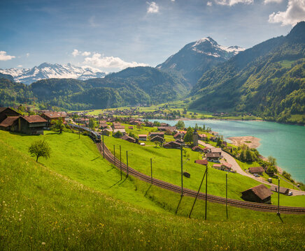 View of the Lungernersee lake with a train passing through the village of Lungern surrounded by lush green meadows and snow-capped mountains in Lungern, Obwalden, Switzerland.
