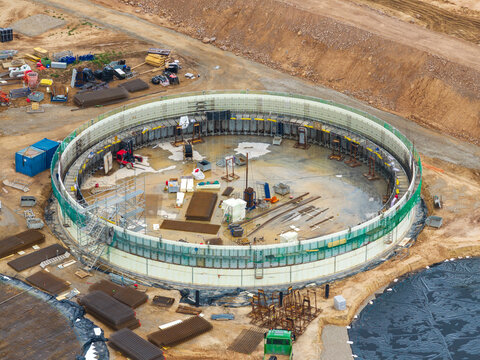 Aerial view of a circular construction site featuring steel reinforcement structures, scaffolding, and heavy machinery on a dirt ground in Lithuania.