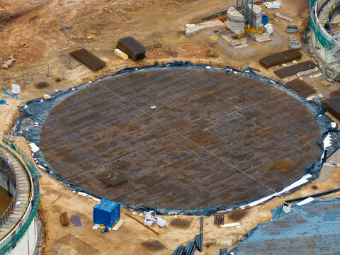 Aerial view of a large circular construction foundation with a steel rebar grid, a blue storage container, and surrounding earthworks in Lithuania.