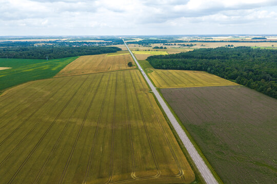 Aerial view of a straight road cutting through vast agricultural fields with golden and green crops, bordered by dense forests under a cloudy sky in Lithuania.
