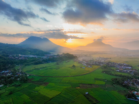 Aerial view of lush green rice fields surrounding a village nestled between mountains during a golden sunrise in Kadungora, West Java, Indonesia.