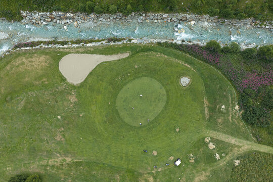 Aerial view of a golf course with a putting green, sand bunkers, and a mountain stream flowing through the alpine landscape in Breuil-Cervinia, Aosta Valley, Italy.