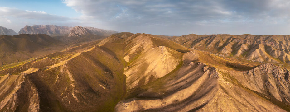 Aerial view of the rugged mountain landscape near Kol-Suu, featuring rolling golden hills, dramatic rocky peaks, and a vast sky in At-Bashi District, Naryn Region, Kyrgyzstan.