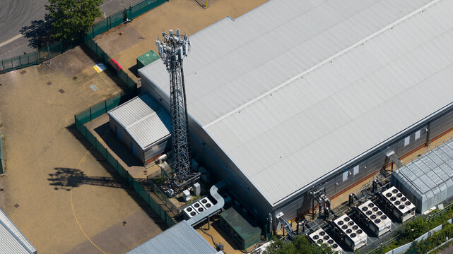 Aerial view of a telecommunications tower situated next to a large industrial facility featuring rooftop HVAC units and fenced perimeter in Hemel Hempstead, England, United Kingdom.
