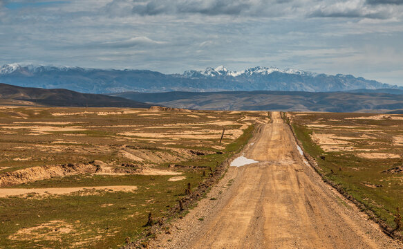 View of a dirt road stretching through the vast arid landscape with snow-capped mountains in the distance under a cloudy sky in At-Bashi District, Naryn Region, Kyrgyzstan.