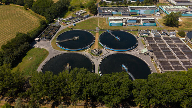 Aerial view of a wastewater treatment plant featuring large circular sedimentation tanks and industrial infrastructure surrounded by lush green trees and fields in Milan, Lombardy, Italy.