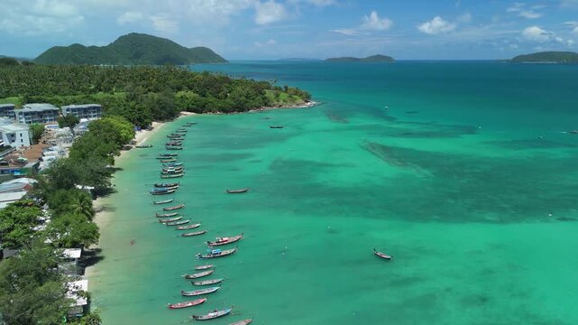 Aerial view of Rawai Beach with traditional longtail boats anchored in turquoise water along the coastline of Tambon Rawai, Chang Wat Phuket, Thailand.