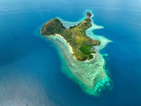 Aerial view of CYC Beach with its white sand shore, lush green vegetation, and vibrant turquoise coral reefs surrounded by the deep blue waters of the ocean in Coron, Mimaropa, Philippines.