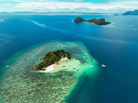 Aerial view of CYC Beach featuring a small island with white sand, surrounded by turquoise waters, coral reefs, and traditional outrigger boats in Coron, Mimaropa, Philippines.