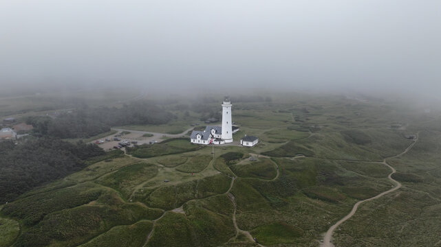 Aerial view of Hirtshals fyr surrounded by grassy dunes and rolling hills under a thick blanket of fog in Hirtshals, Denmark.