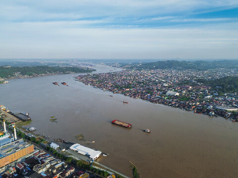 Aerial view of the Mahakam River flowing through the city with coal barges, dense urban housing, and a mosque along the riverbank in Samarinda, Kalimantan Timur, Indonesia.