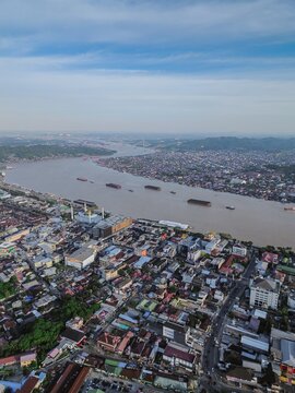 Aerial view of the Mahakam River flowing through Samarinda, featuring dense urban rooftops, a mosque, and cargo barges on the water in Samarinda, Kalimantan Timur, Indonesia.