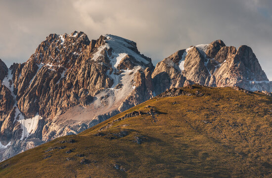 Aerial view of Kol-Suu mountain range featuring rugged peaks with snow patches, golden grassy slopes, and dramatic clouds in At-Bashi District, Naryn Region, Kyrgyzstan.