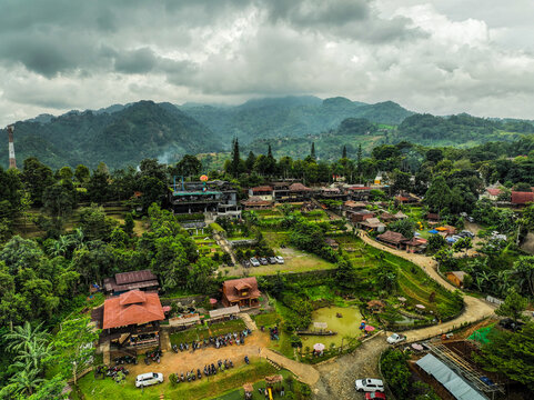 Aerial view of KM 0 Sentul surrounded by lush green mountains, terraced landscapes, and rustic buildings with red roofs in Kecamatan Babakan Madang, Jawa Barat, Indonesia.