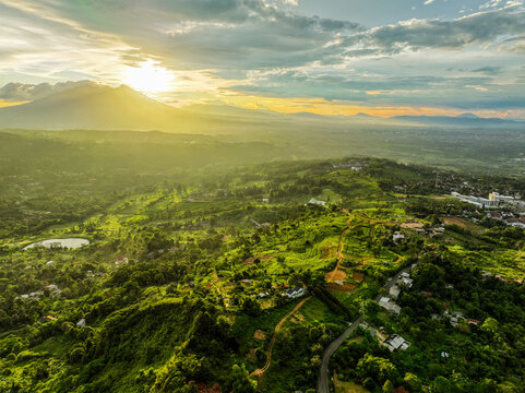 Aerial view of the lush green hills and winding roads surrounding KM 0 Sentul during a golden sunset in Kecamatan Babakan Madang, Jawa Barat, Indonesia.