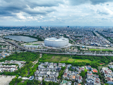 Aerial view of Jakarta International Stadium surrounded by urban sprawl, lush green areas, and a nearby lake in Tanjung Priok, Jakarta, Indonesia.