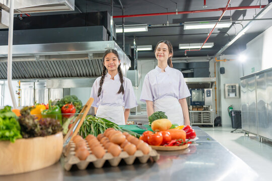 Young asian girls in chef uniform smiling in commercial kitchen ready for cooking class, Child cooks standing behind table full of fresh ingredients in cooking school kitchen